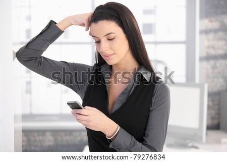 Smiling woman using mobile phone for texting, standing in office, thinking.?