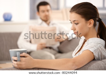 Young attractive woman sitting on sofa at home drinking tea, bored, man reading newspaper in the background.?