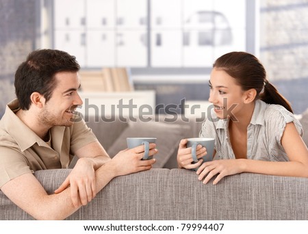 Happy couple sitting on sofa at home, drinking tea, smiling at each other.?