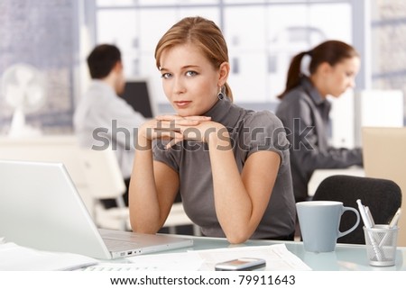 Young female office worker sitting at desk in office, using laptop, colleagues working in background.?