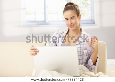 Morning portrait of happy woman having coffee, using laptop computer on sofa, smiling at camera.?