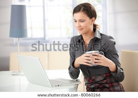 Young female browsing internet at home, drinking tea, smiling.?