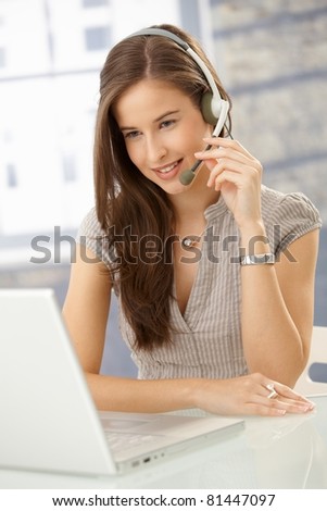 Smiling woman speaking on headset, using laptop computer, looking at screen.