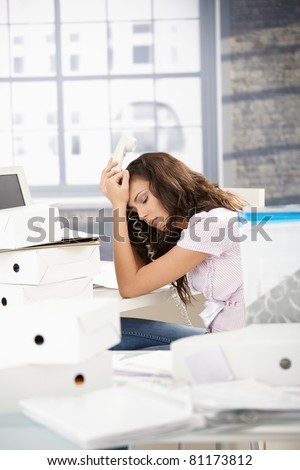 Young girl having headache in office, sitting at desk, holding phone.?