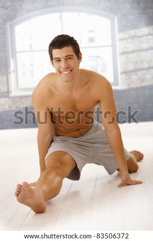 Masculine guy doing forward split in gym, smiling happily at camera.?