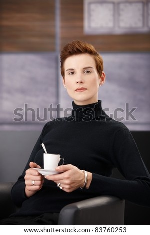 Smart woman wearing polo-neck top having coffee in armchair, holding saucer, looking at camera.?