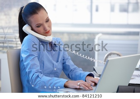 Busy office worker girl taking landline phone call while typing on laptop computer.?