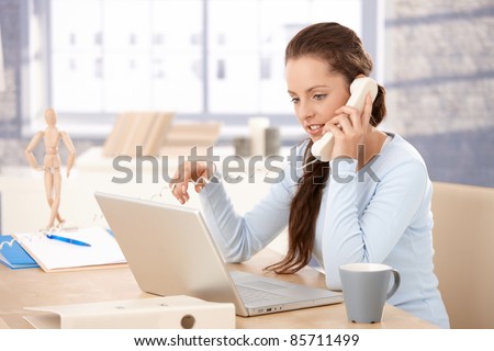 Attractive young girl working on laptop in bright office, talking on phone, looking at screen.?