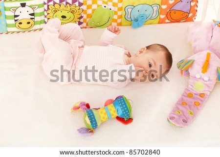 Baby girl looking at colorful toy in bed, with hand in mouth.?