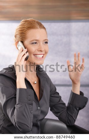 Happy young businesswoman laughing on phone call, gesturing with hand, sitting in office lobby.?