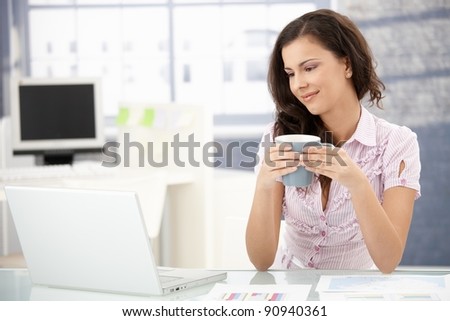 Attractive young woman browsing internet in bright office, drinking tea, smiling.?
