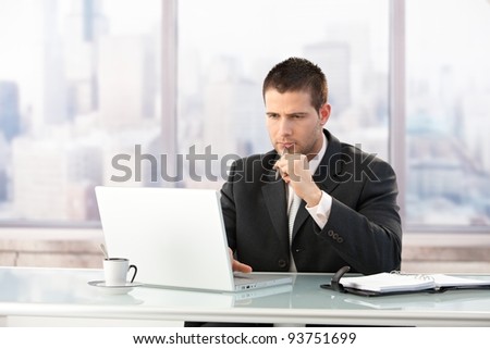 Young manager sitting at desk in bright office, working on laptop.?