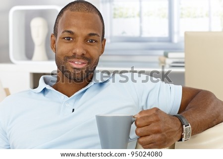 Portrait of cheerful handsome black man sitting at home with mug handheld, drinking tea, smiling at camera.?
