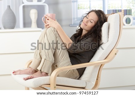 Woman relaxing with closed eyes and cup of tea in armchair at home, daydreaming.