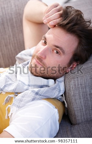 Handsome young man resting at home, laying on sofa, looking at camera.
