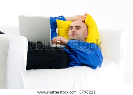 Man in casual business attire, laying on a white couch against a yellow pillow, using a laptop placed on his stomach.  Pleasant expression.  Isolated on white background.