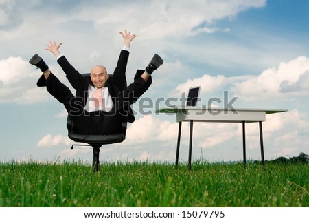 A view of an ecstatic businessman, sitting in a chair in the middle of a grassy field with arms and legs high in the air.
