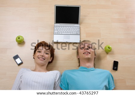 A couple laying on the floor with a laptop, cellular phones and green apples.