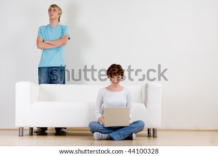 A young couple in a plain white room, near a white couch or sofa.  Woman sitting on floor, using a laptop computer.