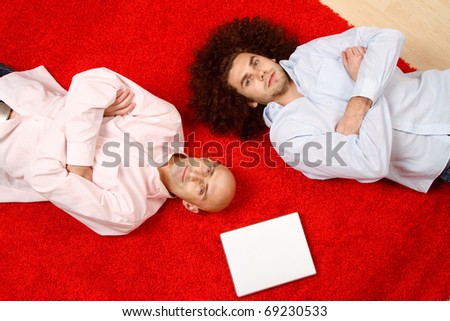 Two men laying side by side on their backs on a red rug in opposite directions, with their arms crossed in front of them.  A blank white paper is on the rug near them.