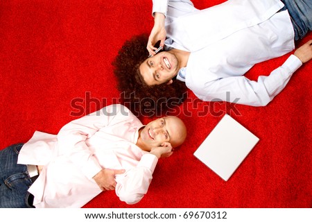 Two men with phones to their ears, laying on their backs in opposite directions on a bright red rug with a pad of paper beside them.  Shot taken from directly above them.