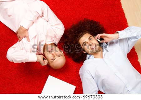 Two men with phones to their ears, laying on their backs in opposite directions on a bright red rug with a pad of paper beside them.  Shot taken from directly above them.