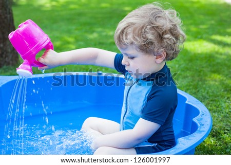 Little blond toddler boy playing with water in summer garden on sunny day