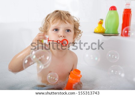 Adorable blond toddler boy playing with soap bubbles by taking bath in bathtub