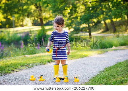 Beautiful little girl of 2 in rain boots playing with yellow rubber ducks in summer park.