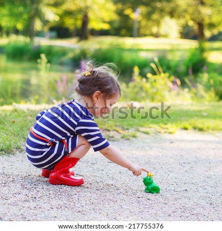Adorable little toddler girl in red rain boots playing with rubber toy frog. Outdoors. Fairytale concept. Square format.