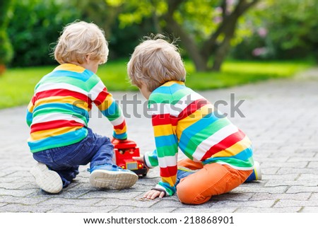 Two siblings, kid boys in colorful clothing with stripes playing with red school bus and toys in summer garden on warm sunny day. Learning to play and communicate together.