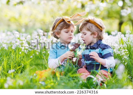 Family of two siblings: Little boys wearing Easter bunny ears at spring green grass and blooming apple garden, eating chocolate bunny and having fun outdoors. On warm sunny day