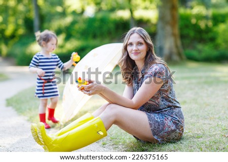 Happy beautiful woman and her little cute kid girl in rain boots, playing together in summer sunny park, on warm day.