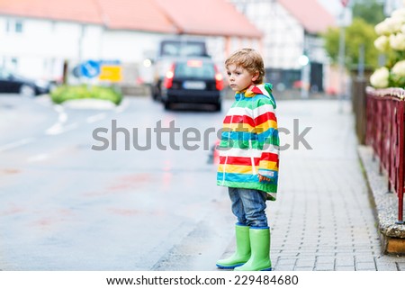 Funny cute kid boy walking in city through rain, wearing colorful rain coat and green boots outdoors at rainy day. Having fun.