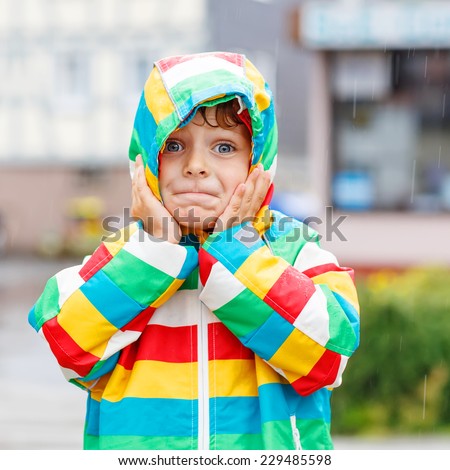 Funny smiling little boy walking in city through rain, wearing colorful rain coat and green boots outdoors at rainy day. Having fun. Square format.