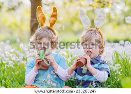 Two little friends with Easter bunny ears at spring green grass and blooming apple garden, eating chocolate bunny and having fun outdoors. On warm sunny day