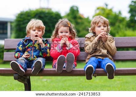 Three children: two little boys and one girl sitting on bench and eating chocolate