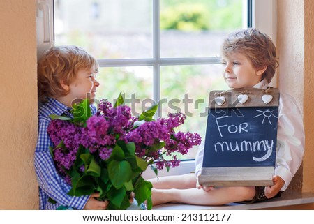 Two happy adorable little sibling boys with blooming lilac flowers and blackboard as gift for their mum on mother's day, indoor. Selective focus on one child.