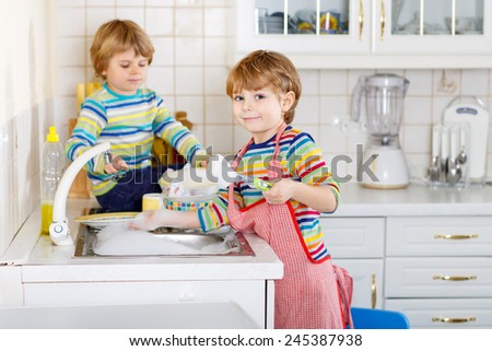 Two funny little boy friends washing dishes in domestic kitchen. Children having fun with helping with housework. Indoors, kids in colorful clothes. Selective focus