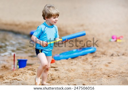 Little blond child having fun with splashing water near city lake, playing water gun. Outdoors leisure with kids in summer, on sunny hot day.