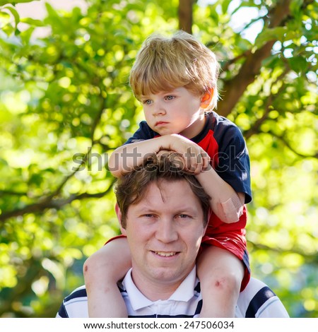 Father carrying child on his shoulders in the park. Little blond kid boy is sad.