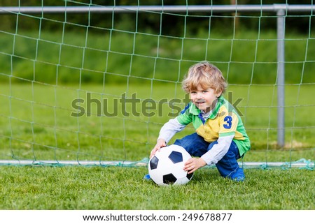 Funny happy little kid boy playing soccer and football and having fun, outdoors on field. Active leisure with children on warm sunny summer day.