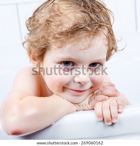 Adorable  blond toddler boy having fun with water by taking bath in bathtub at home. Funny kid splashing and playing with toys.