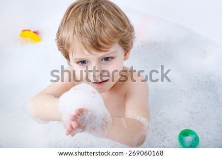 Funny blond toddler boy having fun with water by taking bath in bathtub at home. Cute kid splashing and playing with foam.