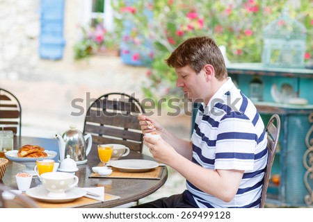 Young man enjoying vegan breakfast outside in summer with various jams, bread, coffee, croissants, muesli and orange juice. French healthy breakfast.