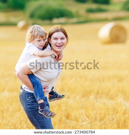 Adorable blond kid boy and his mother having fun on yellow hay field in summer. Happy family of two enjoying nature and togetherness. Boy riding on mum.