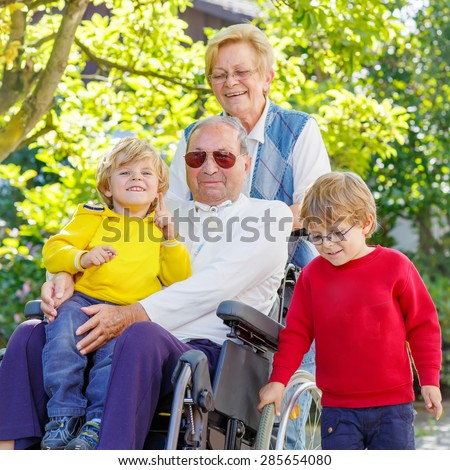 Two little kid boys, their grandmother and grandfather in wheelchair in summer garden. Happy family of four spending time together, outdoors.
