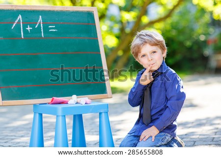 Little funny boy at blackboard thinking about mathematics, outdoor school or nursery. Kid learning and schoolboy concept. On summer sunny day.