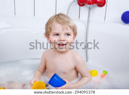 Happy cute little  blond toddler boy having fun with water by taking bath in bathtub at home. Funny kid splashing and playing with toys.