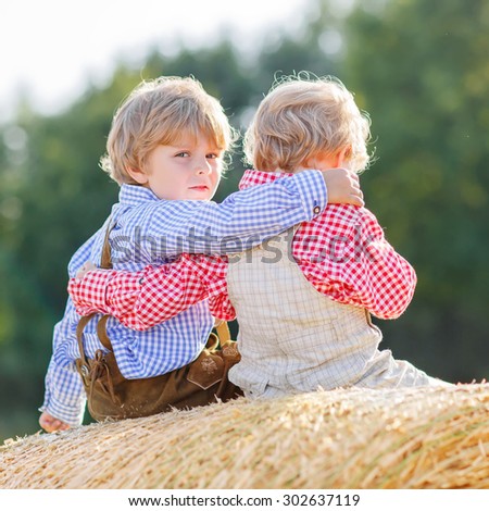 Two little friends sitting on hay stack or bale and speaking on yellow wheat field in summer. Children wearing traditional bavarian clothes.